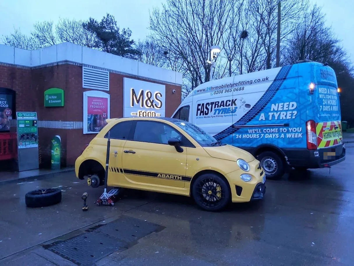 Yellow Abarth 595 having a wheel changed beside the Fastrack van at an M&S forecourt