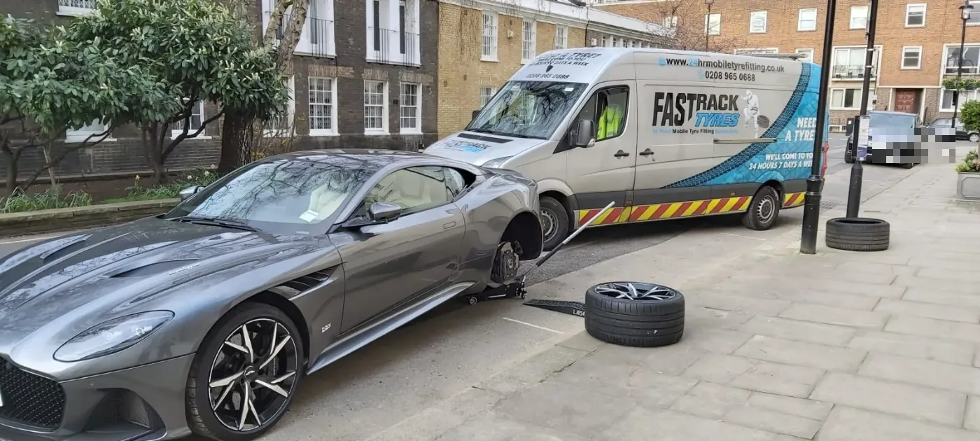 Grey Aston Martin DBS Superleggera raised on a jack for a daytime wheel change beside the Fastrack van