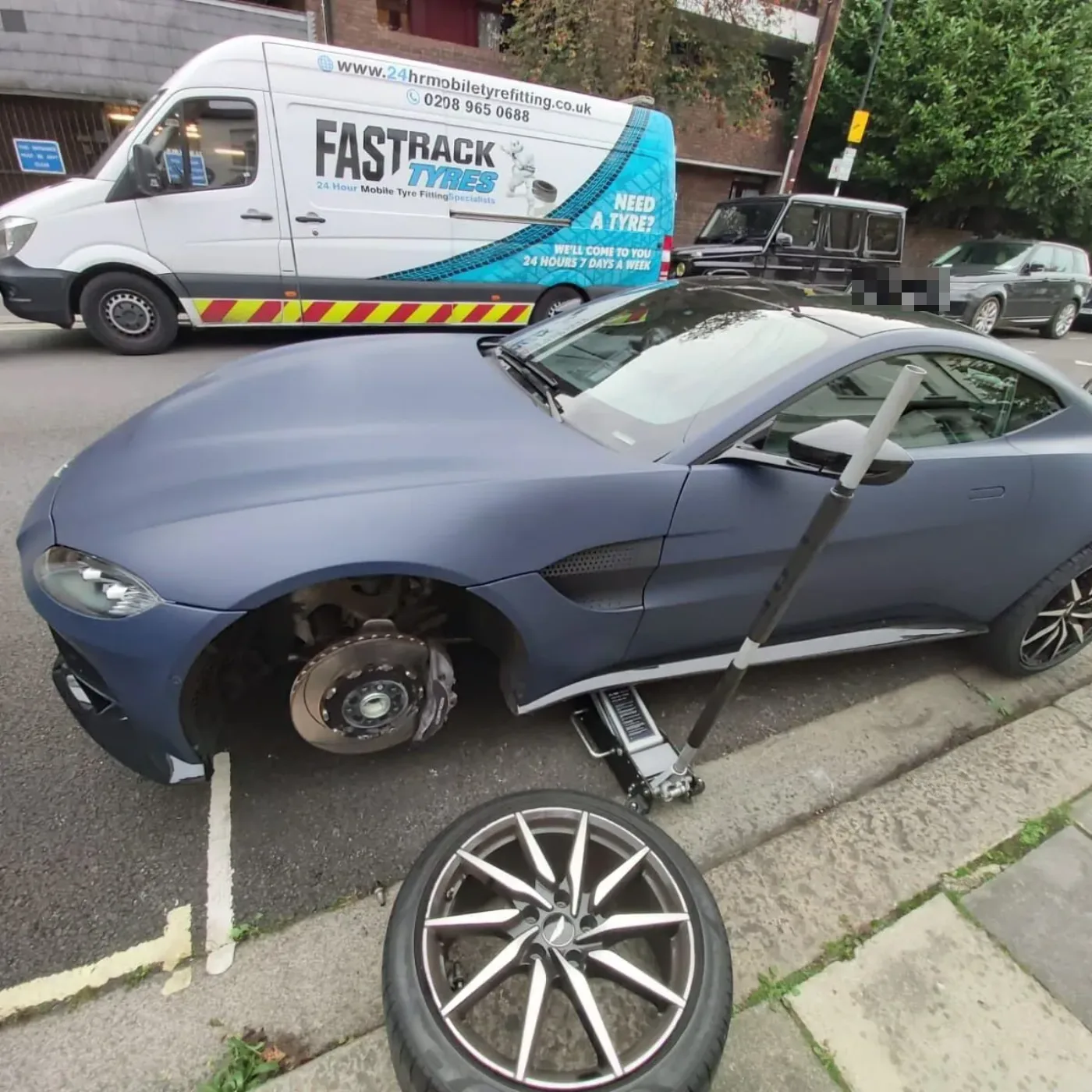 Satin-blue Aston Martin Vantage having a wheel changed on a daytime street beside the Fastrack van