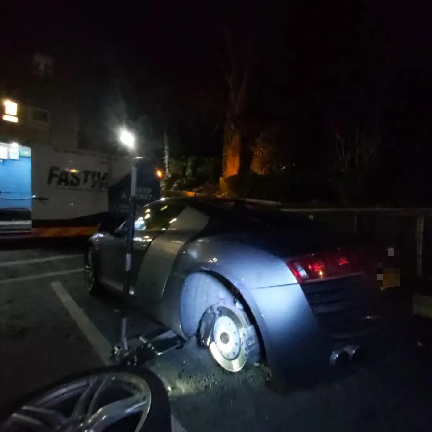 Grey Audi R8 under a streetlight at night with its rear wheel off beside the Fastrack van