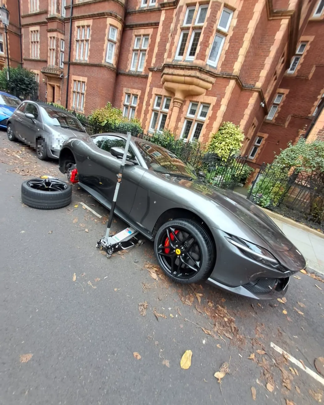 Grey Ferrari Roma on the jack for a wheel-off tyre change outside a mansion block