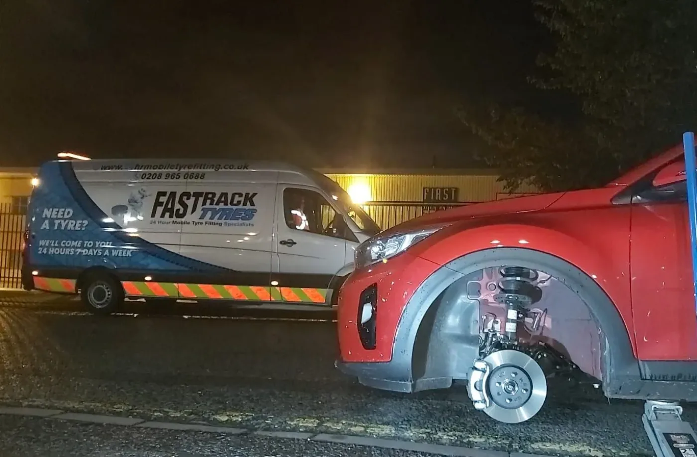 Red Kia having a front wheel changed at night beside the Fastrack van