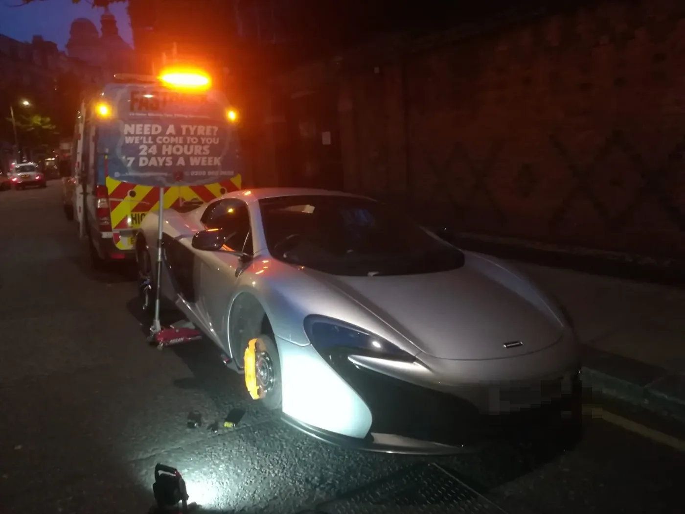 Silver McLaren 570S having a wheel changed at night under amber beacons