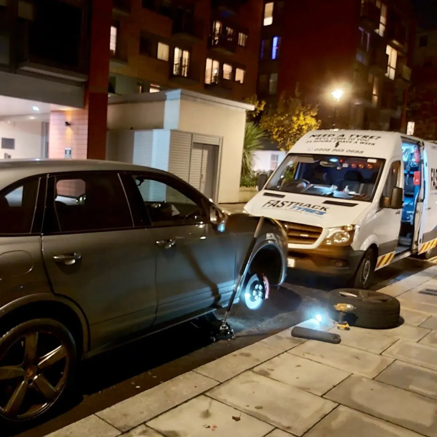 Grey Porsche Cayenne having a wheel changed at night with the spare and tools laid out beside the Fastrack van