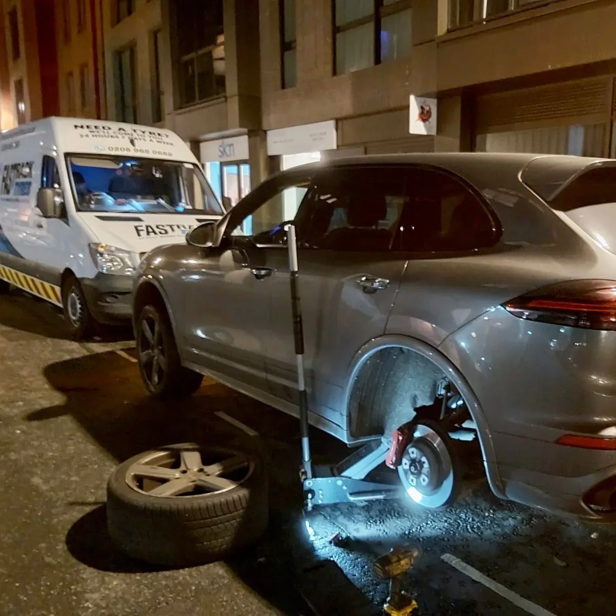 Silver Porsche Cayenne with its rear wheel removed at night and the Fastrack Tyres van behind