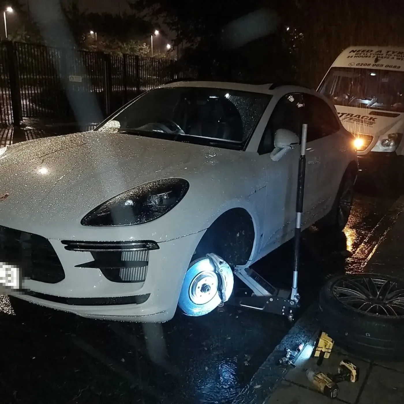 White Porsche Macan having a front wheel changed on a wet night with the spare and power tools laid out