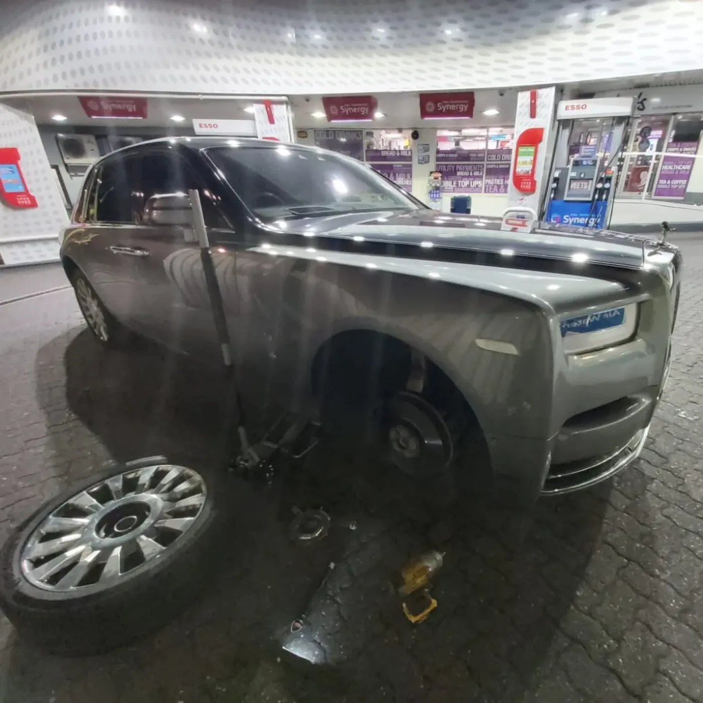 Grey Rolls-Royce Phantom on an Esso forecourt at night with its wheel off and the spare alloy on the ground