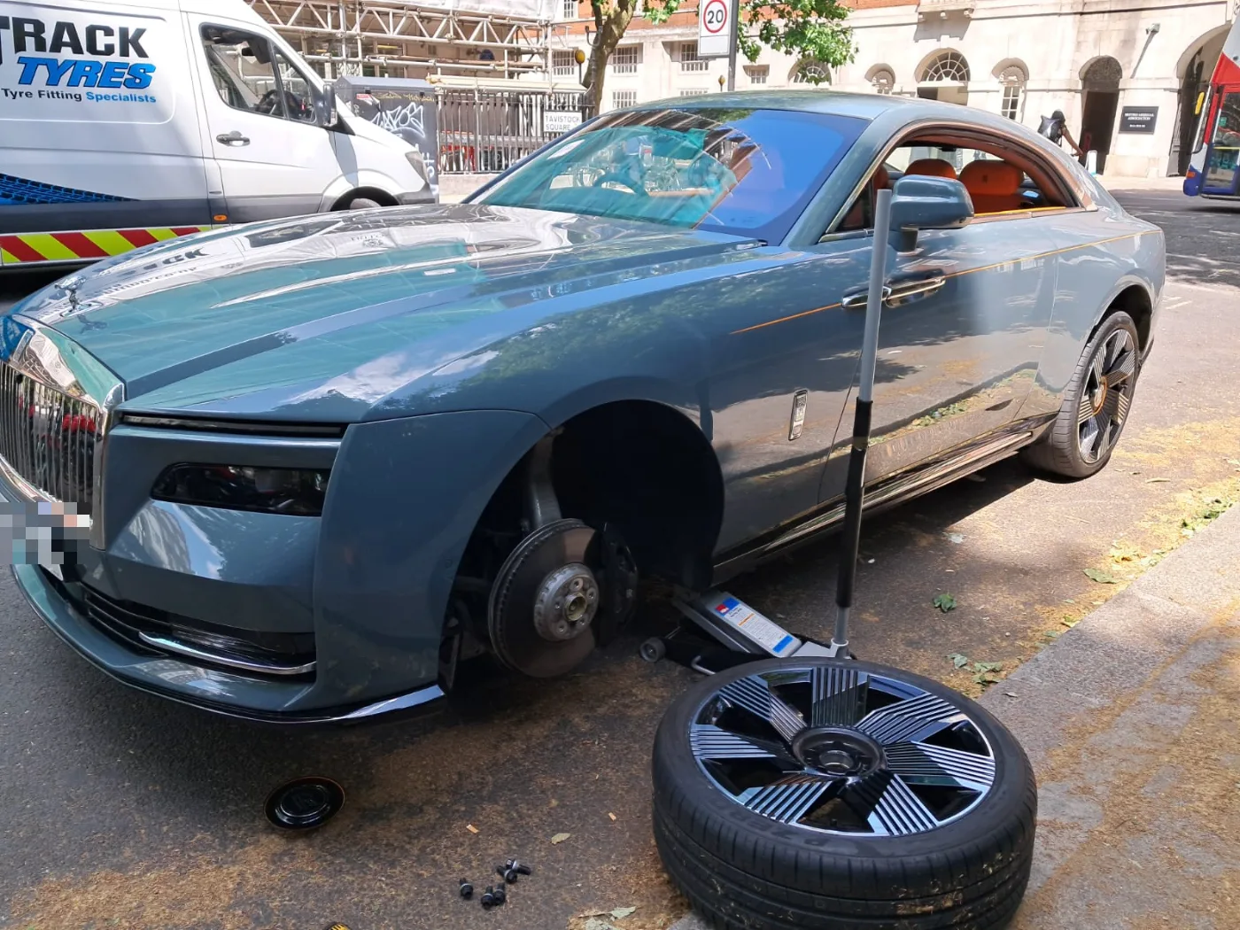 Blue-grey Rolls-Royce Spectre electric grand tourer raised on a trolley jack for a mobile tyre change on a London street, the Fastrack van behind