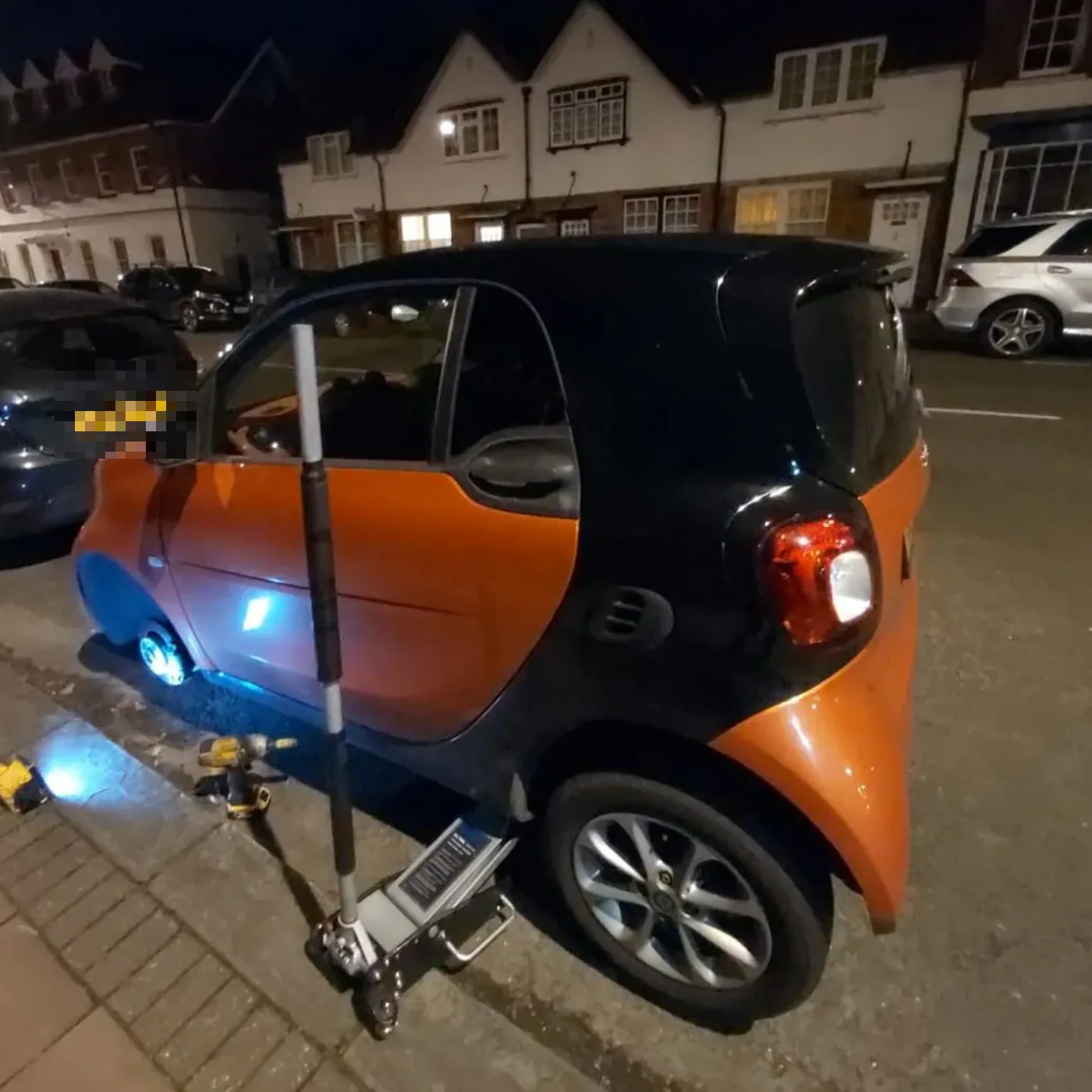 Orange and black Smart ForTwo raised on a trolley jack for a night-time wheel change on a residential street
