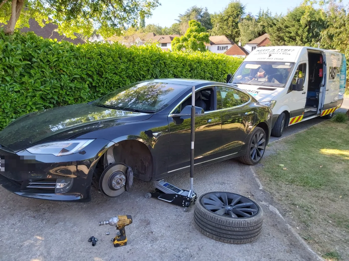 Black Tesla Model S having a tyre replaced on a residential street on a sunny day, Fastrack van alongside